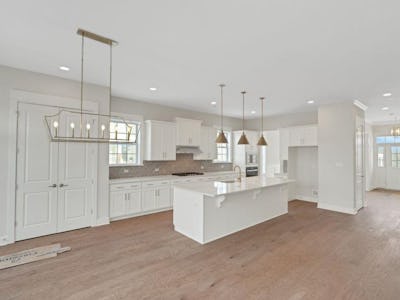 A modern kitchen features white cabinetry, an island with a countertop, pendant lighting, and wooden flooring. The space is well-lit with ample natural light and has a cozy, open layout.